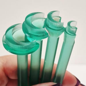 May include: Close-up of several translucent teal-colored plastic tools. Each tool has a long, thin stem and a curved, semi-circular head. The tools are held in a hand with dark purple nail polish.