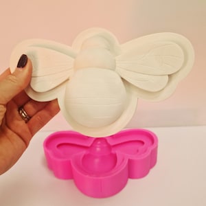 May include: A white bee-shaped mold held by a hand, with a pink base. The bee mold has detailed wing and body features. The pink base has a flower-like shape.