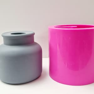 May include: A gray, matte-finish vase with a rounded body and a narrow neck sits next to a bright pink, cylindrical mold. The vase has a classic shape, while the mold is smooth and modern. Both objects are on a white surface.