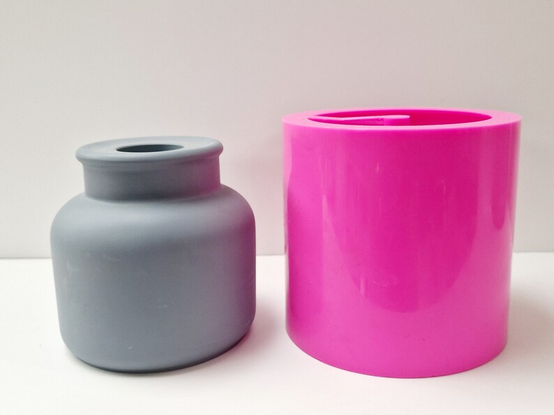 May include: A gray, matte-finish vase with a rounded body and a narrow neck sits next to a bright pink, cylindrical mold. The vase has a classic shape, while the mold is smooth and modern. Both objects are on a white surface.