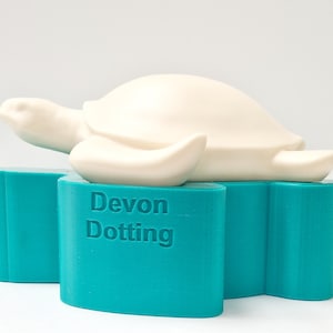 May include: A white ceramic turtle figurine resting on a teal blue base. The base has the text "Devon Dotting" embossed on the front.