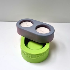 May include: A two-piece silicone mold for making concrete candle holders. The top mold is gray and has two circular depressions for tealight candles. The bottom mold is green and has a single circular depression. The bottom mold is labeled "Devon_Dottie".