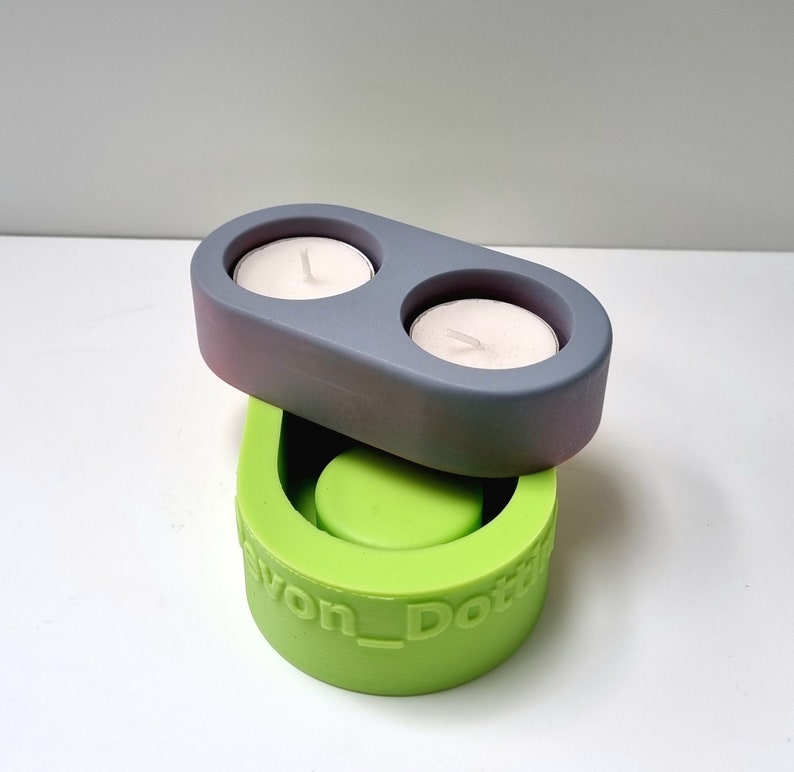 May include: A two-piece silicone mold for making concrete candle holders. The top mold is gray and has two circular depressions for tealight candles. The bottom mold is green and has a single circular depression. The bottom mold is labeled "Devon_Dottie".