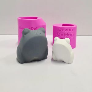 Könnte beinhalten: Zwei froschförmige Objekte, eines grau und eines weiß, stehen vor zwei rosa zylindrischen Behältern. Auf den Behältern steht der Text "Devon Dotting". Die Froschfiguren sind glatt und stilisiert.