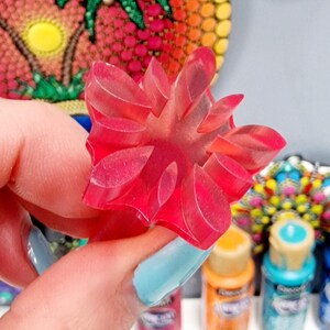 May include: A close-up of a pink plastic flower-shaped stamp. The stamp is being held by a hand with blue nail polish. The stamp is surrounded by paint bottles and white daisy stickers.