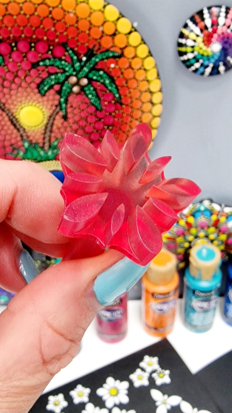 May include: A close-up of a pink plastic flower-shaped stamp. The stamp is being held by a hand with blue nail polish. The stamp is surrounded by paint bottles and white daisy stickers.