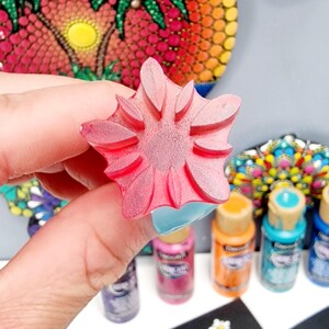 May include: A pink flower-shaped rubber stamp with a raised design. The stamp is being held by a hand. There are several bottles of paint in the background, along with a black surface with white flower designs.