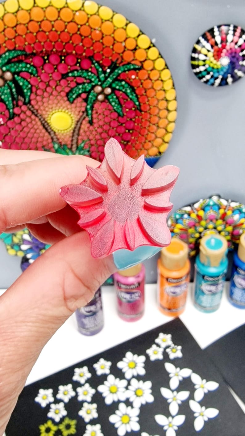 May include: A pink flower-shaped rubber stamp with a raised design. The stamp is being held by a hand. There are several bottles of paint in the background, along with a black surface with white flower designs.