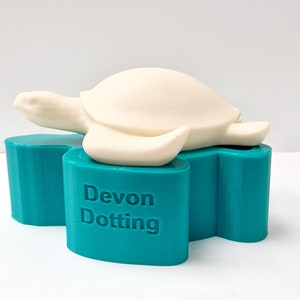 Könnte beinhalten: Eine weiße Keramik-Schildkrötenfigur sitzt auf einem türkisfarbenen Sockel mit dem Aufdruck "Devon Dotting" auf der Vorderseite.