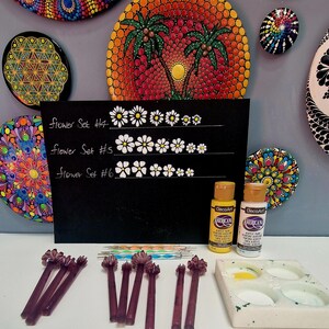 May include: An assortment of mandala art pieces, including painted stones and a black chalkboard with flower set designs. The image also features paint bottles, dotting tools, and a paint palette, suggesting a DIY art project.