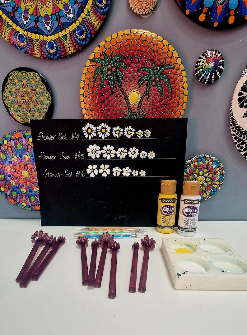 May include: An assortment of mandala art pieces, including painted stones and a black chalkboard with flower set designs. The image also features paint bottles, dotting tools, and a paint palette, suggesting a DIY art project.