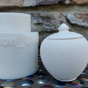 Może przedstawiać: Dwa białe ceramiczne przedmioty, okrągły słoik z pokrywką i cylindryczny pojemnik z wytłoczonym tekstem "Devon_D" na boku.