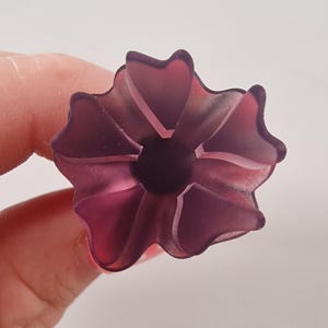 May include: A translucent, purple flower-shaped object with a dark center. The object has multiple petals and is held by a person's fingers against a white background. The flower appears to be made of glass or a similar material.