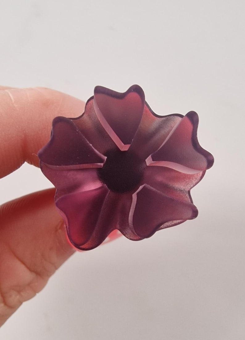 May include: A translucent, purple flower-shaped object with a dark center. The object has multiple petals and is held by a person's fingers against a white background. The flower appears to be made of glass or a similar material.