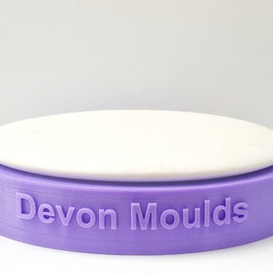 Könnte beinhalten: Eine runde Silikon-Tortenform in Lila mit dem geprägten Text "Devon Moulds" an der Seite. Die Form hat einen abnehmbaren weißen Boden.