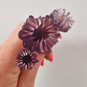 Op de afbeelding: Drie paarse, bloemvormige objecten met ingewikkelde, radiale ontwerpen. De objecten hebben een doorschijnende kwaliteit, met een donkerder centrum en lichtere, bloembladachtige randen. De bloemen worden tegen een effen witte achtergrond gehouden, wat hun delicate structuur laat zien.
