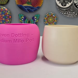 Puede incluir: Dos pequeñas macetas redondeadas. Una es de color rosa vibrante con el texto "Devon Dotting - Medium Milo Pot". La otra es de color blanco crema. Ambas tienen un acabado liso y mate. Obras de arte decorativas en el fondo.