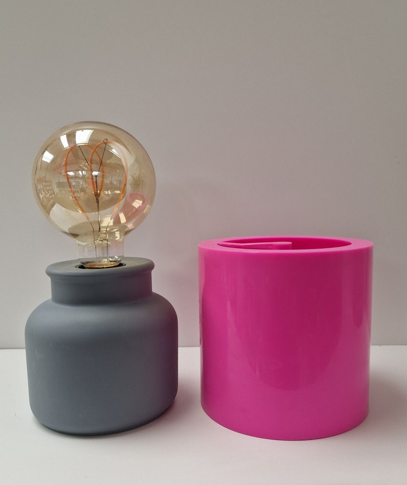 May include: A gray, ceramic lamp base with a large, round, Edison-style light bulb. Next to it is a bright pink, cylindrical container. The lamp has a classic, minimalist design. The bulb has a warm, amber glow.