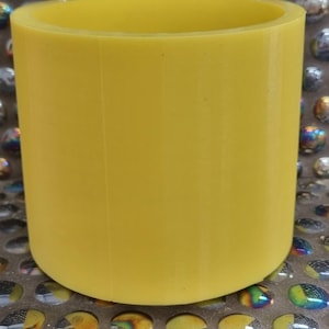 May include: A bright yellow cylindrical container with a smooth, slightly textured surface. The container is empty and appears to be made of plastic.