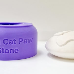 May include: A purple silicone mold for making a 10cm cat paw stone. The mold is next to a white cat paw stone.