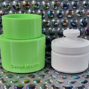 Peut inclure: Un moule à bougie en plastique vert en deux parties. Le moule est cylindrique et possède un dessus amovible. Le fond du moule porte l'inscription "Devon Moulds". À côté du moule se trouve un pot à bougie en céramique blanche avec un couvercle.