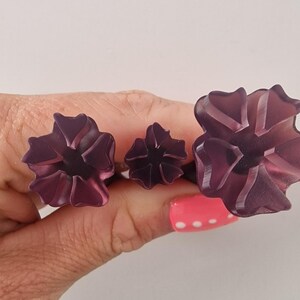 May include: Three dark purple flower-shaped rings. The rings are held in a hand with pink nail polish. The flowers have a translucent appearance with detailed petal designs. The rings are likely fashion accessories.