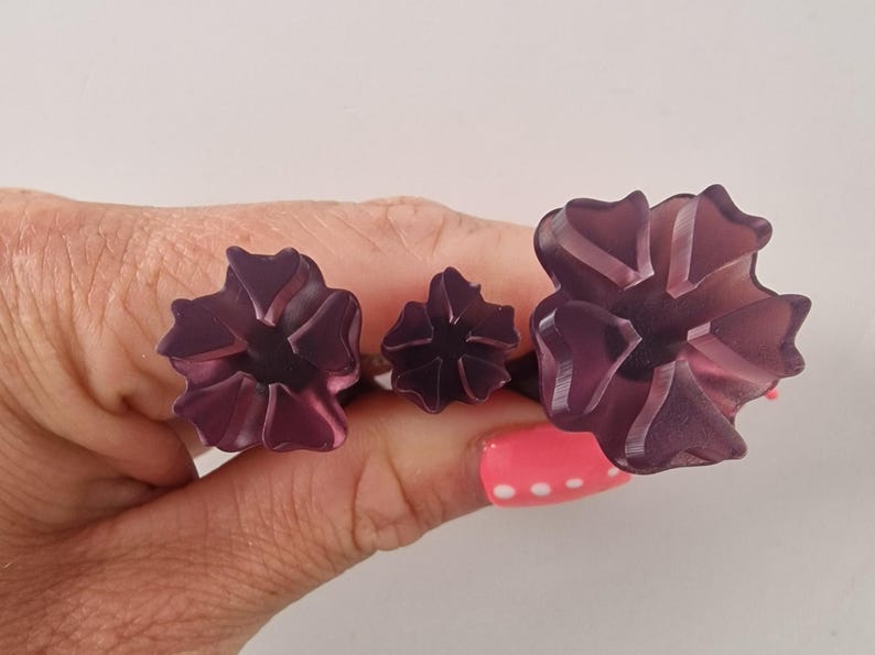 May include: Three dark purple flower-shaped rings. The rings are held in a hand with pink nail polish. The flowers have a translucent appearance with detailed petal designs. The rings are likely fashion accessories.