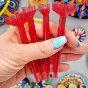 May include: Four red plastic tools with a unique design, used for creating dot art. The tools are held in a hand with light blue nail polish. The tools are in front of a row of paint bottles with various colors.