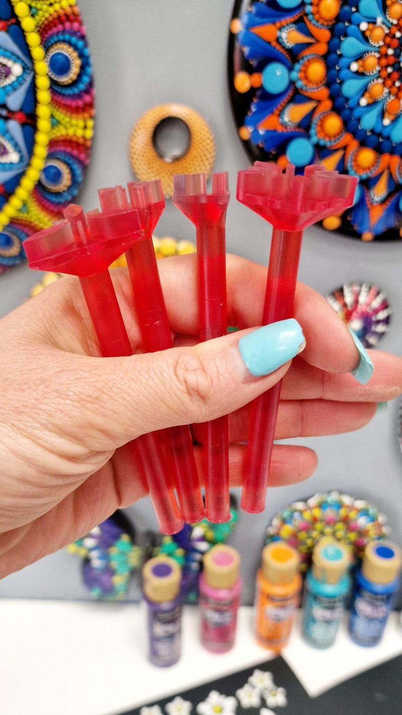 May include: Four red plastic tools with a unique design, used for creating dot art. The tools are held in a hand with light blue nail polish. The tools are in front of a row of paint bottles with various colors.