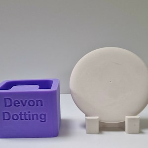 Könnte beinhalten: Eine violette Silikonform zum Backen eines runden Kuchens oder Desserts. Die Form trägt die Aufschrift "Devon Dotting" auf der Vorderseite. Ein runder Kuchenständer aus weißer Keramik mit einem kleinen Loch in der Mitte ist ebenfalls abgebildet.