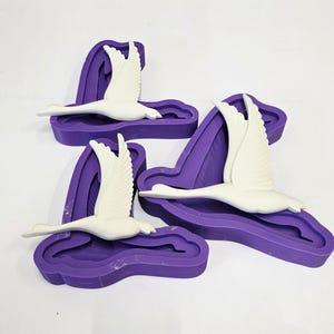 May include: Three white swan figurines in flight, each set within a deep purple silicone mold. The swans have outstretched wings and are positioned at different angles. The molds have a wavy, organic shape.