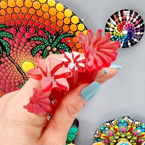 Può includere: Un set di tre strumenti di punteggiatura in plastica rossa per creare l'arte del mandala. Gli strumenti hanno la forma di fiori e hanno punti di diverse dimensioni. Gli strumenti sono tenuti in una mano con smalto per unghie blu chiaro.