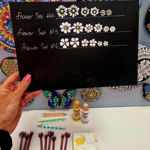 May include: A black chalkboard displays three flower designs labeled "Flower Set #4", "Flower Set #5", and "Flower Set #6". The image also shows painting tools, paint bottles, and a butterfly-shaped decorative item.