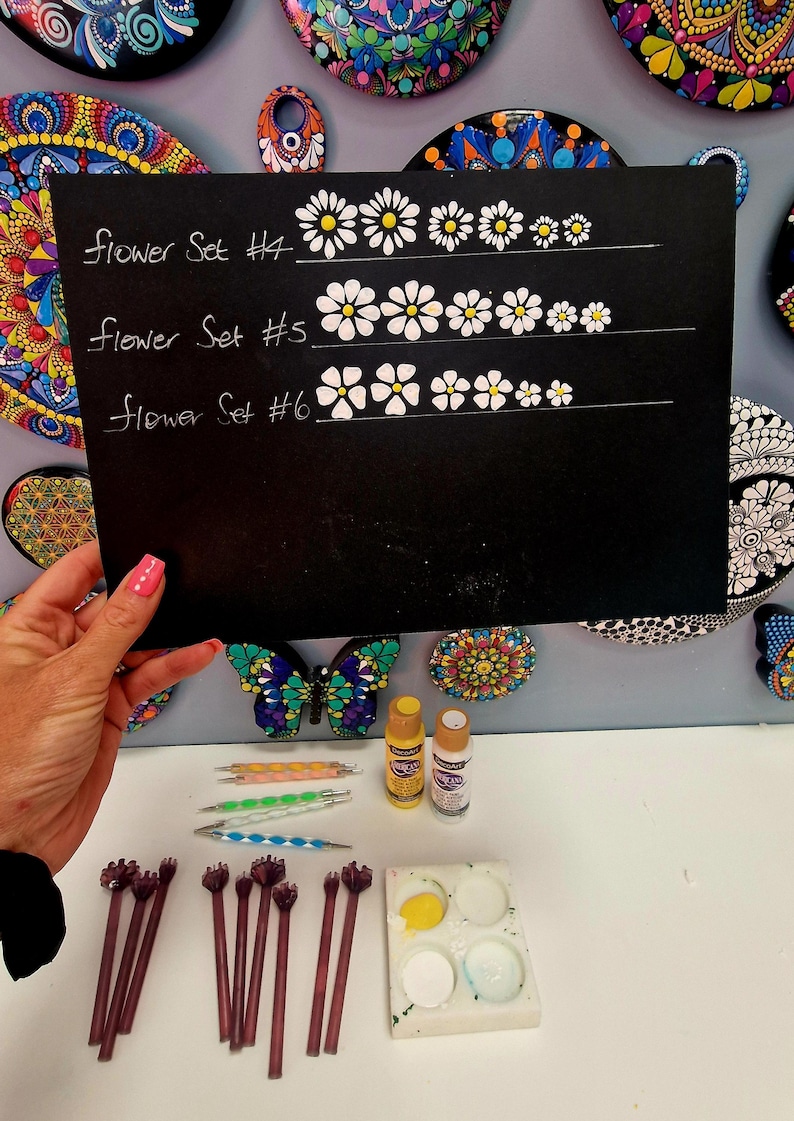 May include: A black chalkboard displays three flower designs labeled "Flower Set #4", "Flower Set #5", and "Flower Set #6". The image also shows painting tools, paint bottles, and a butterfly-shaped decorative item.