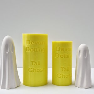 Könnte beinhalten: Drei gelbe Kerzen mit dem Text "Devon Dotting - Tall Ghost" darauf. Zwei weiße Geisterfiguren stehen links und rechts neben den Kerzen.