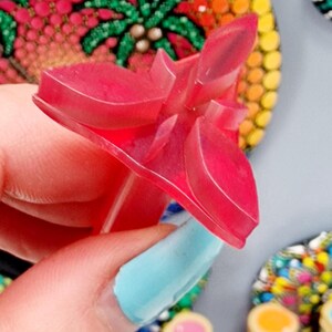 May include: A pink plastic flower-shaped tool for creating dot art. The tool has a pointed tip and a handle. The tool is being held by a hand with blue nail polish.