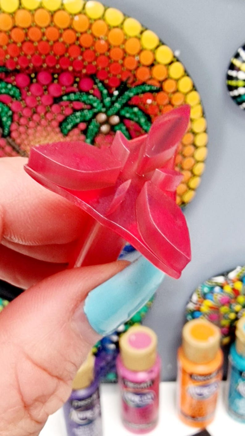 May include: A pink plastic flower-shaped tool for creating dot art. The tool has a pointed tip and a handle. The tool is being held by a hand with blue nail polish.