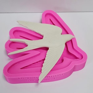 Könnte beinhalten: Eine weiße Schwalbenfigur liegt in einer leuchtend rosa Silikonform. Die Form hat eine detaillierte Kontur der Vogelform mit Text am Boden. Der Vogel hat ausgebreitete Flügel und ein detailliertes Federdesign.