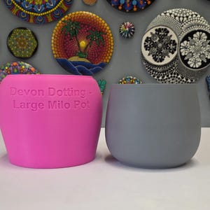 Könnte beinhalten: Zwei kleine, abgerundete Pflanzgefäße. Eines ist leuchtend rosa, das andere ein gedämpftes Grau. Auf dem rosa Pflanzgefäß steht der Text "Devon Dotting - Large Milo Pot". Diese Pflanzgefäße sind ideal für kleine Pflanzen oder Sukkulenten.