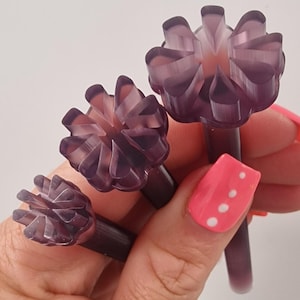 Puede incluir: Tres objetos morados con forma de flor y tallos se sostienen en una mano. Los objetos tienen un diseño de pétalos en capas y un aspecto translúcido. La mano tiene esmalte de uñas rosa.