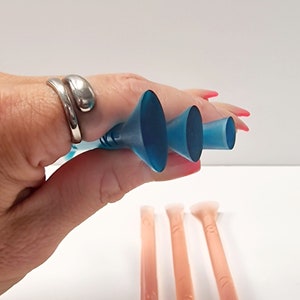 Puede incluir: Tres aplicadores de plástico azul con puntas redondeadas, dispuestos en orden descendente de tamaño, sostenidos en una mano. Tres aplicadores adicionales con puntas redondeadas se muestran en primer plano, en color rosa claro.