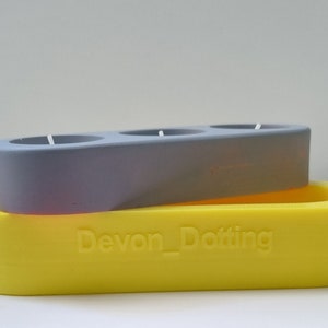 May include: A gray candle holder with three circular openings sits on top of a yellow oval-shaped candle holder. The yellow candle holder has the text "Devon_Dotting" embossed on the front.