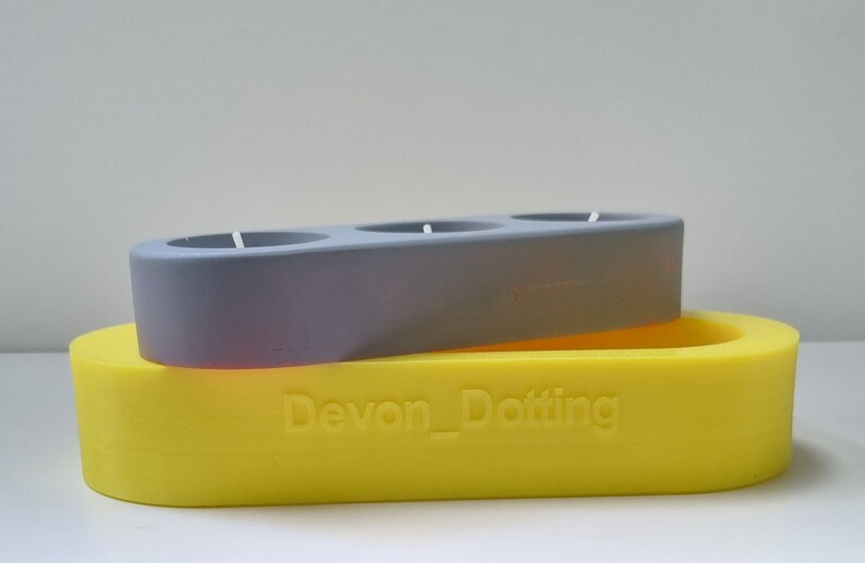 May include: A gray candle holder with three circular openings sits on top of a yellow oval-shaped candle holder. The yellow candle holder has the text "Devon_Dotting" embossed on the front.