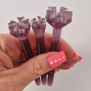 May include: Three translucent purple floral-shaped tools are held in a hand. Each tool has a long stem and a flower-like top with multiple petal-like structures. The tools are a deep purple color.