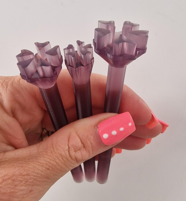 May include: Three translucent purple floral-shaped tools are held in a hand. Each tool has a long stem and a flower-like top with multiple petal-like structures. The tools are a deep purple color.