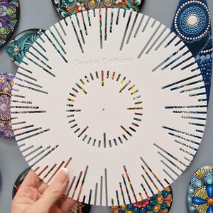 Puede incluir: Un esténcil circular blanco con el texto "Devon Dotting" se sostiene frente a arte de puntos colorido. El esténcil tiene cortes radiales y un agujero central. Las piezas de arte de puntos presentan patrones intrincados y colores vibrantes.