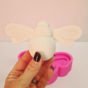 May include: A white, three-dimensional bee figurine with detailed wings and a segmented body. The bee is held in a hand, with a pink mold in the background. The bee is a craft item.