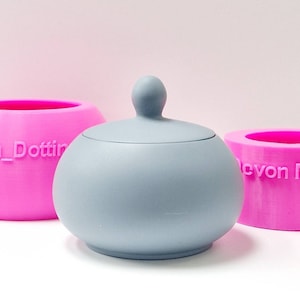 Op de afbeelding: Twee roze siliconen mallen met de tekst "Devon_Dottir" en "Devon Moulds" erop. Een grijze keramische container met een deksel bevindt zich in het midden.