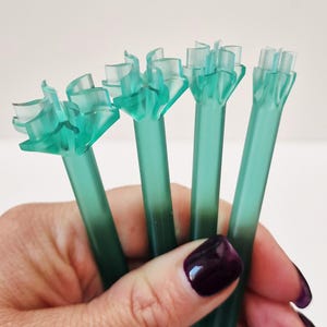 Peut inclure: Ensemble de cinq supports de tiges de fleurs en plastique vert translucide. Chaque support de tige a un corps long et fin avec un dessus évasé à plusieurs pétales. La main a du vernis à ongles violet foncé.