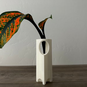 May include: White ceramic vase with a geometric design and a circular opening. The vase is holding a single stem with a large, green and orange leaf.
